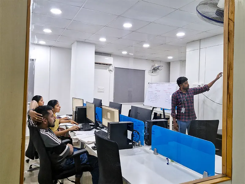 A classroom training session at Cognidel is in progress. A trainer is explaining plc programming concepts on the whiteboard to students. The setting includes multiple desktop systems, a whiteboard with notes, and a focused learning environment.